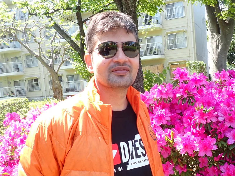 Dr. Ram C. Sharma wearing sunglasses beside bright pink flowering shrubs.